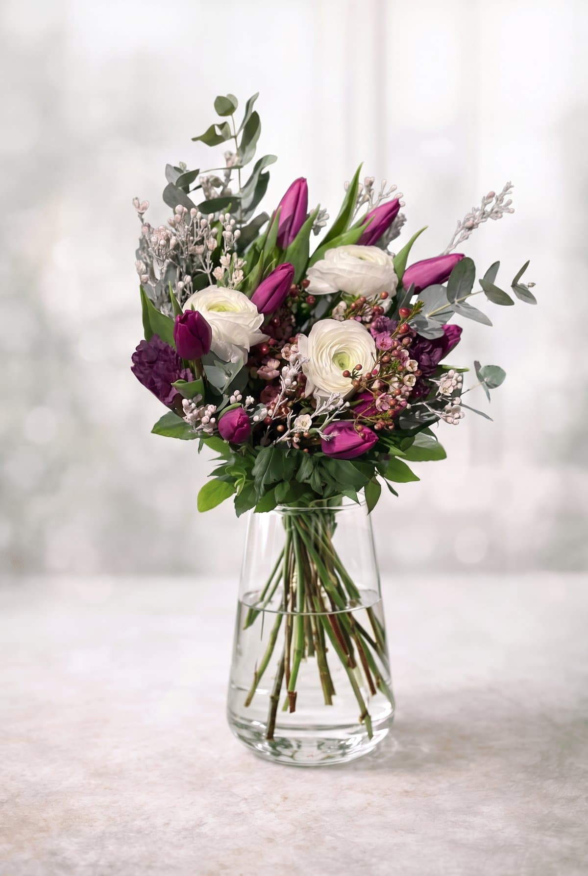 Bukett med lila tulpaner, vita ranunkler, rosa vaxblommor och eukalyptus i glasvas